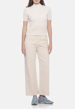 Donna che indossa una maglietta bianca a maniche corte con coste, pantaloni beige chiaro a gamba larga e sneaker azzurre, in piedi davanti a uno sfondo semplice.