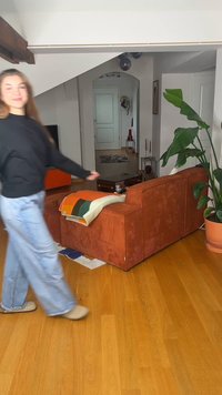 Γυναίκα με μαύρο πουλόβερ και τζιν περπατά δίπλα από πορτοκαλί καναπέ με πολύχρωμη κουβέρτα σε φωτεινό σαλόνι με ξύλινο πάτωμα και γλαστράκι.