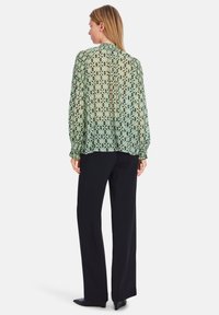 Transparente Bluse mit langen Ärmeln in Hellgrün mit geometrischem Muster, gerafften Manschetten und hohem Kragen; kombiniert mit schwarzen weit geschnittenen Hosen.
