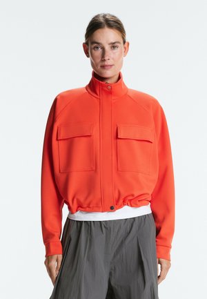 Žena so zviazanými vlasmi v jasne oranžovej bunde s dvoma veľkými prednými vreckami stojaca na bielom pozadí.