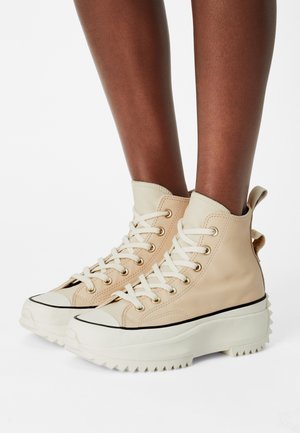 Beige high-top sneakers made of smooth leather, featuring white laces, gold eyelets, and textured rubber soles with a jagged edge.