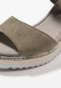 Olivgrön mockaskandal med en bred rem, texturerad plattform, silverfärgade pärlaccenter och sågad vit sula.