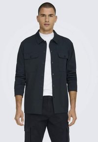 Dunkelblaues Hemd zum Knöpfen mit zwei Brusttaschen, langen Ärmeln, die bis zu den Ellbogen hochgekrempelt sind, und einem strukturierten Kragen, aus einem glatten Stoff gefertigt.