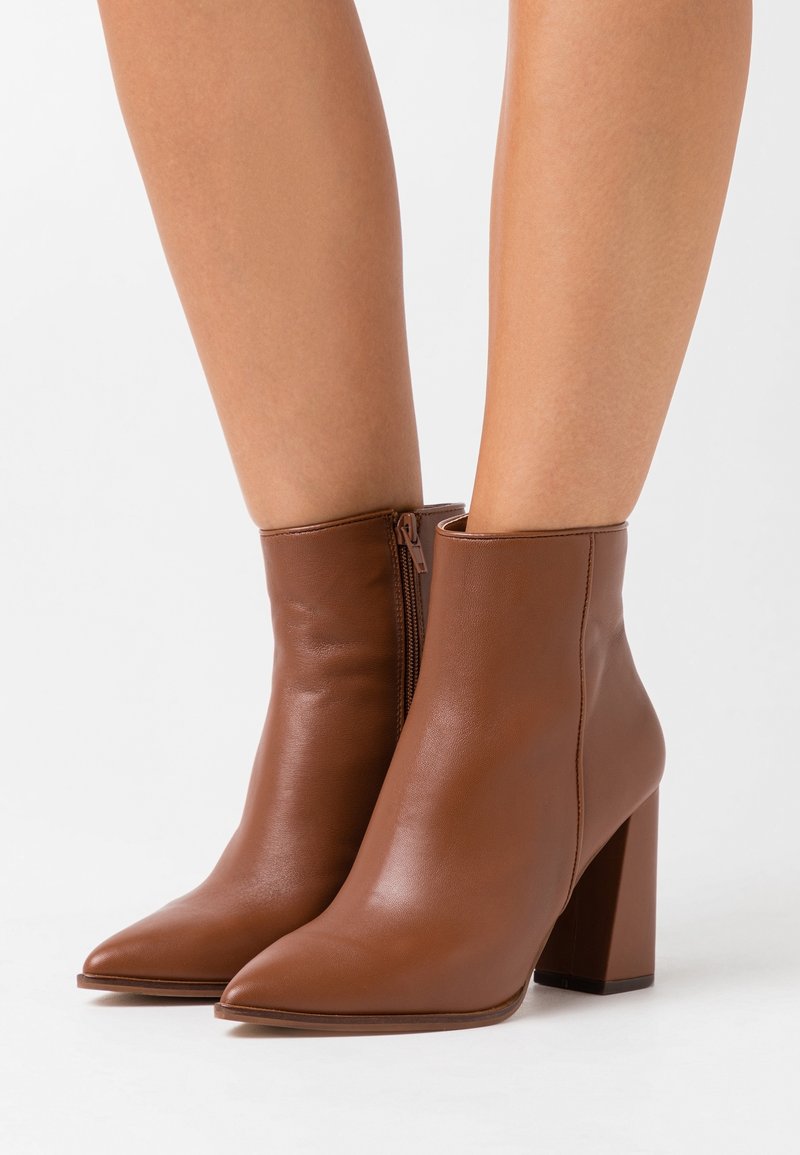 Bottines en cuir marron avec bouts pointus et talons carrés portées par une personne debout devant un fond blanc.