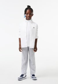 Camicia bianca con colletto sopra un dolcevita bianco, pantaloni sportivi grigi e scarpe da ginnastica blu/bianco. Presenta capelli intrecciati con accenti di perline blu.