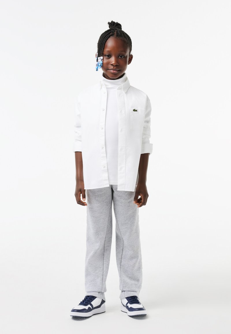 Camicia bianca con colletto sopra un dolcevita bianco, pantaloni sportivi grigi e scarpe da ginnastica blu/bianco. Presenta capelli intrecciati con accenti di perline blu.