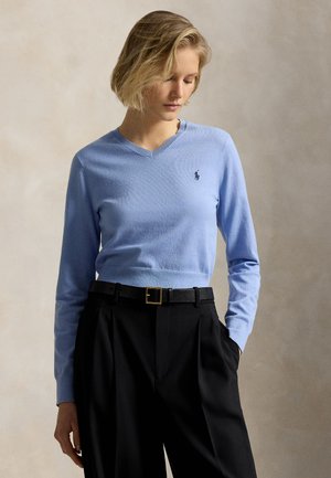 Femme portant un pull bleu clair à col en V avec un petit logo et un pantalon noir taille haute avec ceinture, debout devant un fond uni.