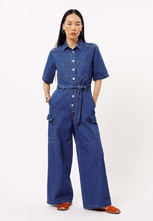 Combinaison en denim à manches courtes, avec deux poches avant et une taille ceinturée. Présente des coutures légères et des jambes larges et amples.