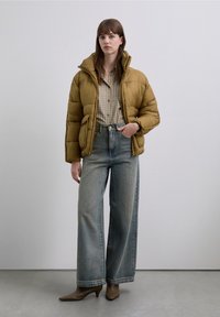 Geacă tip puf de culoare galben muștar, cu guler înalt și design matlasat, combinată cu blugi largi, de culoare albastru deschis, și cizme maro cu toc ascuțit.