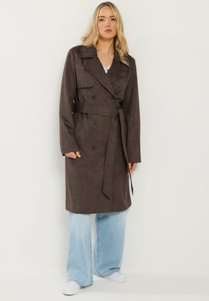 Blonde Frau trägt einen dunkelbraunen Trenchcoat mit Gürtel, hellblaue Jeans mit weitem Bein und weiße Turnschuhe und steht vor einem schlichten Hintergrund.