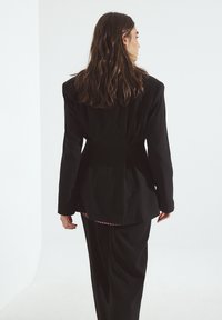 Črn stilsko krojen suknjič z oblikovanimi rameni, oprijetim pasom in gladko teksturo, nosi se čez karo srajco. Enobarvna ozadje.