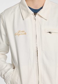 White zip-up jacket made from cotton with a collar. Features "Guess Originals" embroidered in gold, and a zippered chest pocket on the right.