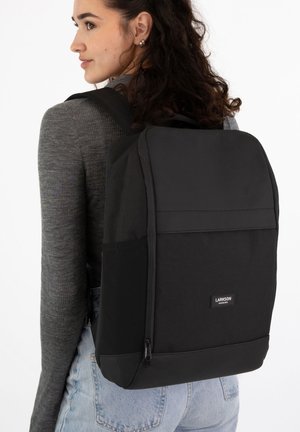 Woman with curly hair wearing a gray sweater and light jeans carries a large black Larkson backpack on her back.