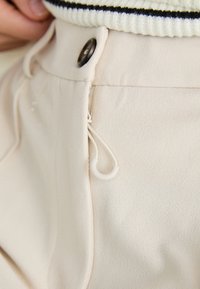 Beige bukser med en glat tekstur, knaplukning og en snøre-sløjfe detalje på taljen.