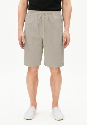 Mann trägt hellbeige knielange Shorts mit Fronttaschen, schwarzes Oberteil und weiße Slipper, steht vor weißem Hintergrund.