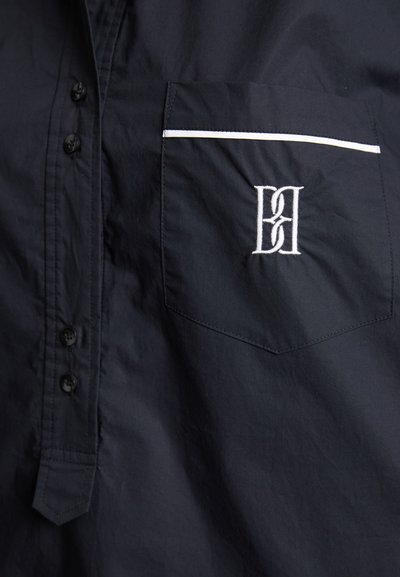 Tumes sinise värvi nööbiga särk, millel on rinnatasku, valge äärestus ja broderi logo. Valmistatud siledast, kergest kangast.