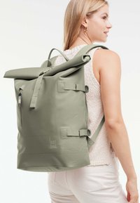 Zelený batoh s rolovacím uzávěrem vyrobený z odolného materiálu, s přední kapsou na zip, nastavitelnými bočními popruhy a s logem na dolní přední části.