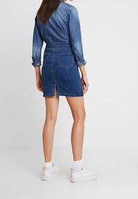 Frau in einem Denimhemd und einem hochgeschnittenen Denimrock mit Rückenschlitz, weißen Socken und weißen Turnschuhen, die an einer schlichten Wand steht.