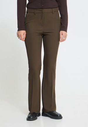 Personne portant un pantalon marron sur mesure, une chemise sombre à manches longues, des chaussures noires et des bagues, debout devant un fond blanc uni.