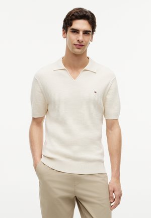 Νεαρός άνδρας στέκεται με τα χέρια στις τσέπες, φορώντας λευκή πλεκτή κοντομάνικη πόλο μπλούζα και μπεζ παντελόνι, μπροστά σε ανοιχτό φόντο.