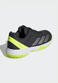 Chaussure de sport noire avec tige en mesh, accents gris métallique, contrefort de talon jaune vif et semelle blanche. Présente un design léger pour une utilisation sur le court.