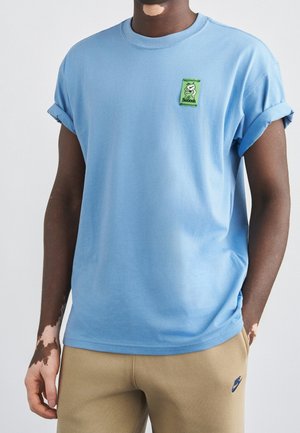 Osoba o ciemnej karnacji, ubrana w niebieską koszulkę z zieloną naszywką graficzną oraz beżowe spodnie dresowe z logo Nike, stojąca z jedną ręką w kieszeni.