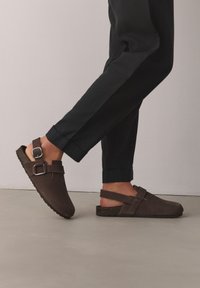 Personne portant des sandales marron foncé fermées à bout fermé avec des brides réglables, associées à un pantalon noir à revers, debout sur un sol clair.