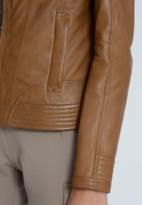 Veste en cuir marron au design ajusté, avec une poche zippée verticale et des détails côtelés le long de l'ourlet et des poignets. Texture lisse.