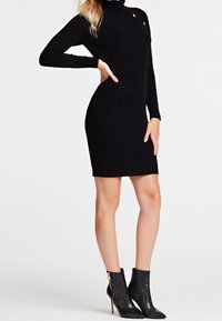 Donna che indossa un vestito a tubino nero a costine con maniche lunghe e collo a imbuto, abbinato a stivaletti neri con punta affilata e dettagli in oro sulla cerniera.