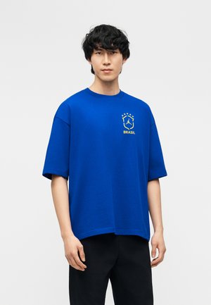 Jonge man met zwart haar die een losse blauwe t-shirt met geel Brazil-logo en zwarte broek draagt, staand tegen een effen lichte achtergrond.