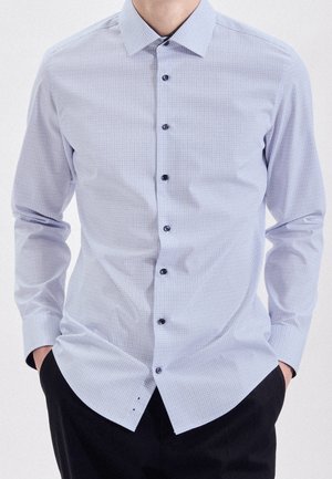 Mann trägt ein hellblaues langärmeliges Hemd mit feinem Karomuster, Hände in den Taschen einer schwarzen Hose.
