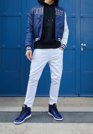 Personne portant une veste universitaire bleu marine et blanche, un sweat à capuche noir, un pantalon blanc et des baskets montantes bleu et noir, debout devant une porte bleue.