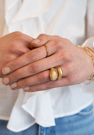 Hände, die ineinander verschränkt sind, tragen zwei goldene Ringe und ein goldenes Kettenarmband, vor einem Hintergrund aus weißer Bluse und blauen Jeans.