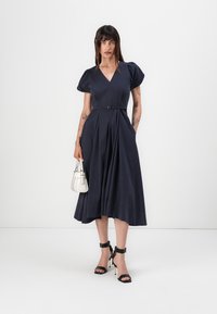 Knielanges navy-blaues Kleid mit Puffärmel, plissiertem Rock und tailliertem Schnitt. Begleitet von einer kleinen weißen Handtasche und schwarzen High Heels.