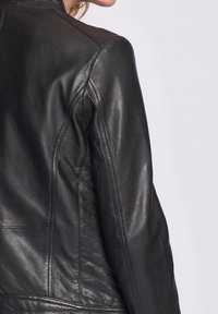 Částečný pohled na osobu v černé kožené bundě, která ukazuje podrobné šití a texturu na zádech a rukávu.