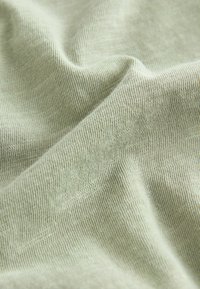 Gaiši zaļa auduma materiāls ar gludu tekstūru, kurā redzami smalki ribojumi. Materiāls izskatās mīksts un tam ir smalkas tonālas variācijas.