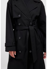 Il trench coat nero presenta un design doppiopetto, un ampio colletto, un dettaglio con cintura in vita e polsini con bottoni, realizzato in un tessuto testurizzato.