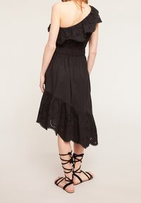 Frau in einem schwarzen, einseitigen Midikleid mit Rüschensaum und Stickerei, kombiniert mit schwarzen schnürbaren flachen Sandalen, stehend vor einem schlichten Hintergrund.