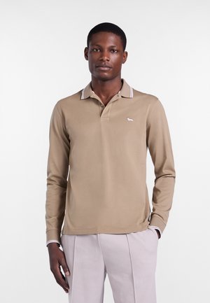 Muškarac u bež polo majici dugih rukava s bijelim malim logotipom psa i svijetlim hlačama stoji s jednom rukom u džepu.