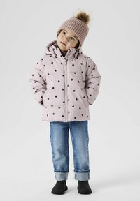 Giubbotto imbottito rosa chiaro con motivi a cuore neri, chiusura con zip, abbinato a jeans in denim blu e stivali neri alla caviglia. Cappello di maglia con pom-pom.