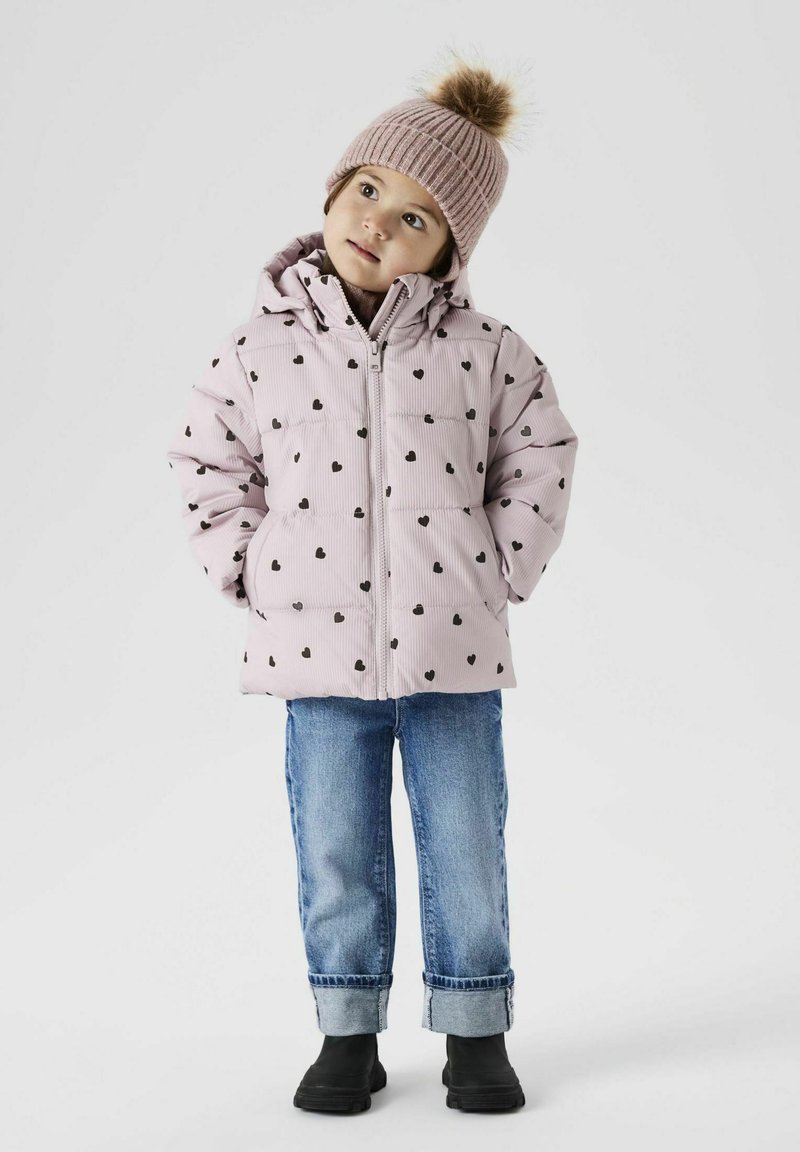 Giubbotto imbottito rosa chiaro con motivi a cuore neri, chiusura con zip, abbinato a jeans in denim blu e stivali neri alla caviglia. Cappello di maglia con pom-pom.