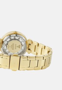 Zlaté kovové náramkové hodinky s okrúhlym puzdrom, priehľadným dnom a prepojeným náramkom. Obsahujú reliéfne akcenty a lesklý povrch.