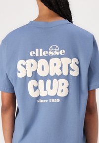 Hele helesinises puuvillases T-särgis on suur valge tekst "ellesse SPORTS CLUB", mille kohal on logo ja allpool tekst "since 1959".