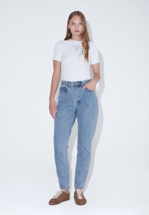 Ανοιχτό μπλε τζιν ψηλόμεσο με ευθεία γραμμή, με κούμπωμα με κουμπιά και διακριτικά σημάδια φθοράς. Συνδυάζεται με εφαρμοστό λευκό μπλουζάκι.