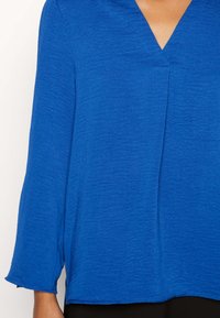 Niebieska bluzka o fakturowanej powierzchni, z dekoltem w kształcie V i rękawami długości 3/4, charakteryzująca się luźnym krojem i miękkim materiałem. Rąbek jest lekko zaokrąglony.