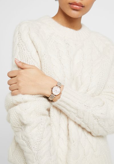 Person iført en hvid chunky kabelstrikket sweater med armene krydset, der viser et sølv- og rosaguld armbåndsur med en hvid urskive.