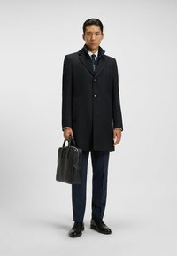 Cappotto nero dal taglio sartoriale, con colletto, chiusura a bottoni monopetto e tasche laterali. Abbinato a pantaloni blu navy e una borsa in pelle.