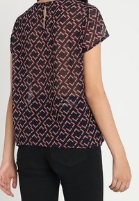 Blouse navy à manches courtes en voile avec un motif géométrique rouge et blanc, dotée d'une ouverture en forme de clé au dos et d'une coupe décontractée.