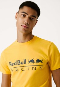 Dzeltena kokvilnas T-krekls ar "Red Bull Racing" tekstu tumši zilā krāsā, zem kura ir grafika ar diviem skrienošu buļļu attēliem. Ar īsām piedurknēm, apaļu kakla izgriezumu.
