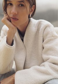 Giacca in pile bianca con superficie testurizzata, maniche ampie e un bordo sottile. Il modello appoggia il mento sulla mano, evidenziando uno sfondo neutro.